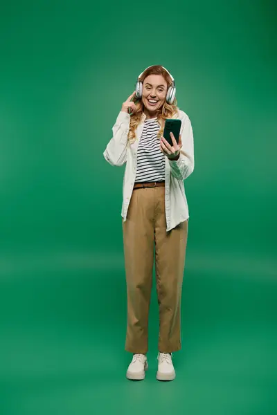 stock image A cheerful woman listens to music with headphones while standing against a vibrant green background.