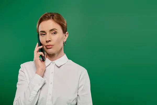 stock image A stylish woman in formal wear speaks on the phone, showcasing professionalism against a green backdrop.