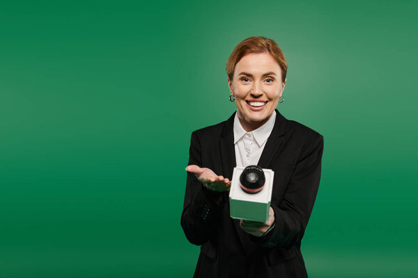 Харизматичный телеведущий в черном вечернем костюме привлекает зрителей на ярком зеленом фоне.