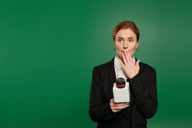 Siyah giyimli şık bir sunucu canlı yeşil arka planda bir mikrofonla şakayla etkileşime girer..