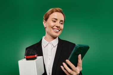 Siyah formalı dengeli bir sunucu elinde akıllı telefon ve mikrofon tutuyor..