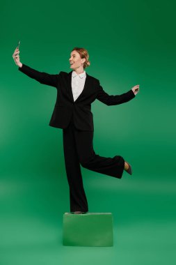 A stylish TV presenter in black formal attire interacts playfully while standing on a pedestal, surrounded by green. clipart