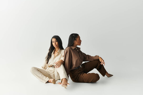 Две молодые женщины излучают уверенность в стильных нарядах, сидя спиной к спине на мягкой поверхности.