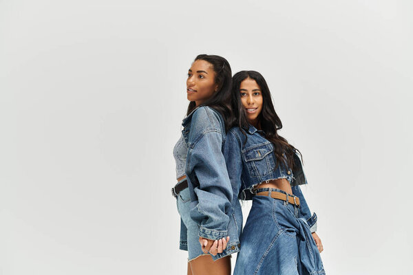 Two young women stand back to back, exuding confidence in trendy denim attire.