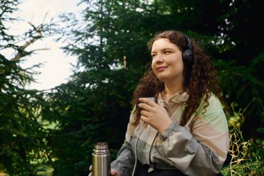 A plus size woman sits in a lush green forest, savoring her drink while listening to music. clipart