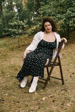 A plus size woman with curly hair relaxes on a chair in a serene field adorned with greenery. clipart