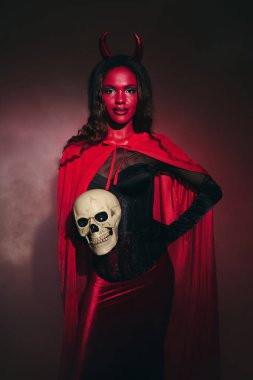 A beautifully dressed woman in a red Halloween costume holds a skull, striking a fierce pose. clipart