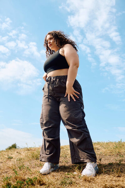 A joyful plus size woman stands tall in a field, embracing nature under a bright sky.