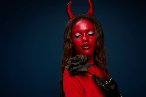 The captivating woman in red devil attire holds a dark rose, embodying Halloween spirit.