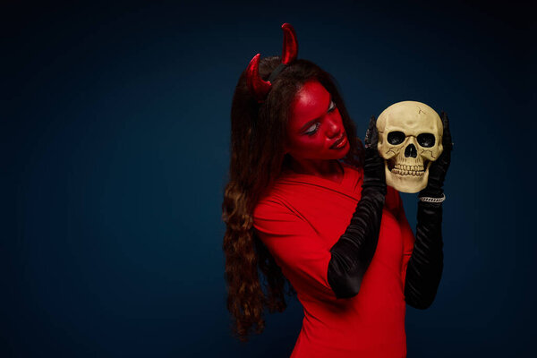 A stunning woman in a devil outfit passionately examines a skull, embracing Halloween spirit.