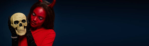Красивая женщина, одетая в костюм дьявола, восхищается черепом в темном капризном наряде на Хэллоуин.