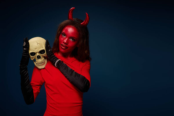 A captivating woman dressed as a devil playfully poses with a skull, embodying Halloween spirit.
