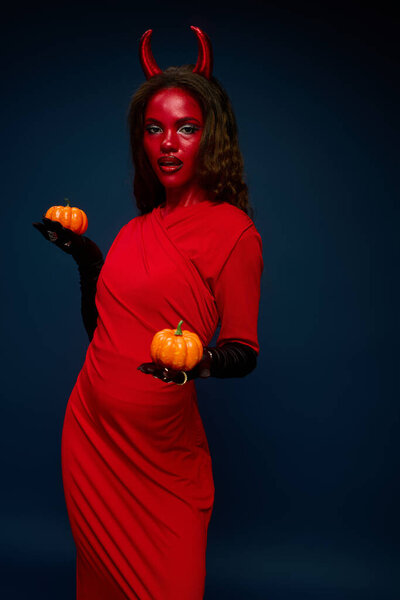 A captivating woman dressed in a devil costume holds small pumpkins, embodying the Halloween spirit.