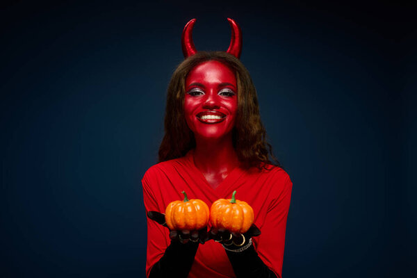 A striking woman in a devil costume beams while holding two small pumpkins, celebrating Halloween.