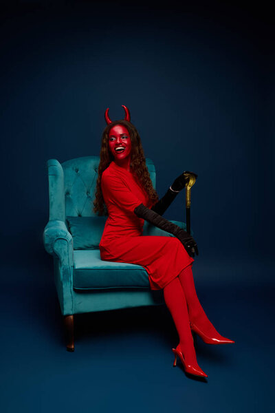 A stunning woman in a bright red devil costume sits confidently in a vintage chair.