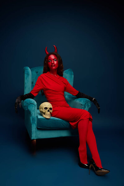 Glamorous woman in a striking red costume and horns sits elegantly beside a decorative skull.