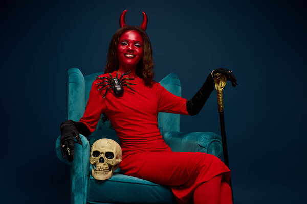 Dressed in a vibrant red outfit and devil horns, she exudes confidence, holding a cane.