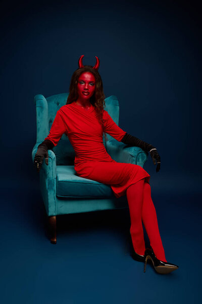 A striking woman dressed as a devil lounges in a teal chair, exuding Halloween spirit.