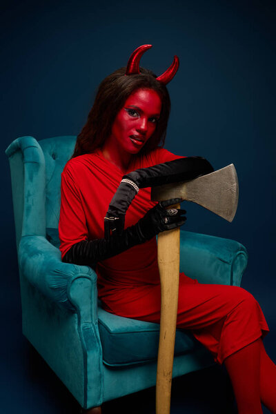 A captivating woman dressed as a devil lounges in a plush blue chair, holding an axe.