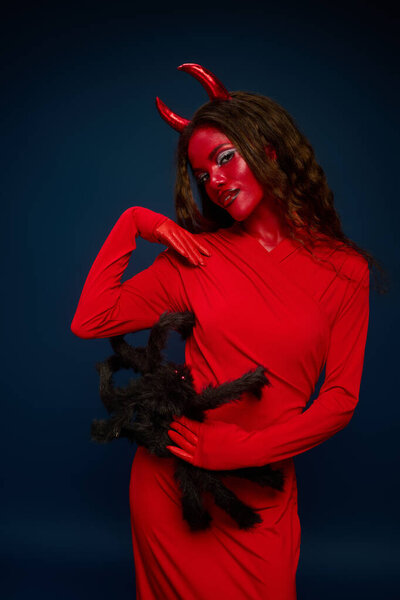 A captivating woman in red playfully shows a black spider, embodying Halloween spirit.