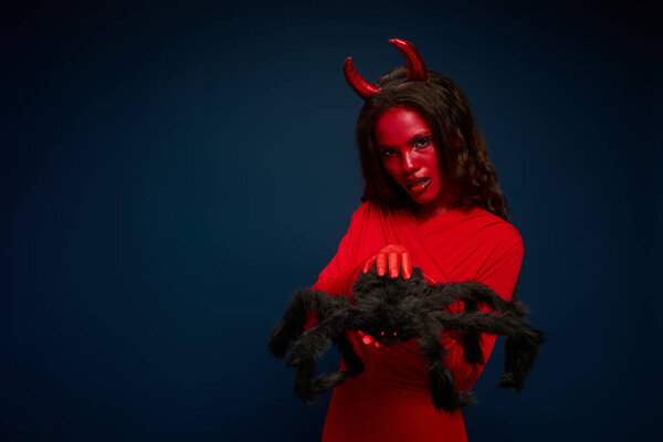 A captivating woman dressed as a devil holds a giant spider, embodying Halloween spirit.