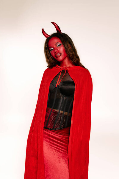 A stunning woman in a red cape and devil horns embraces Halloween at a festive event.