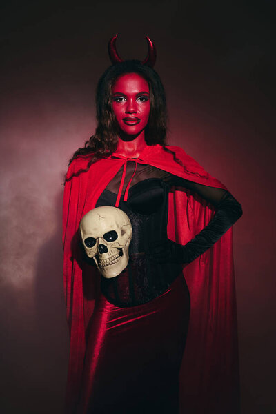 A beautifully dressed woman in a red Halloween costume holds a skull, striking a fierce pose.