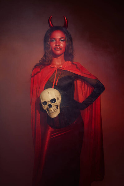 A striking woman dressed as a devil poses confidently, showcasing her Halloween spirit with a skull.