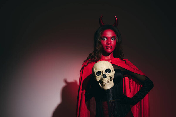 Dressed as a devil, she confidently holds a skull, celebrating the spirit of Halloween.