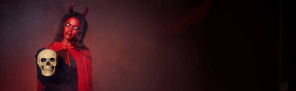 A stunning woman dressed as a devil stands confidently, holding a skull, celebrating Halloween.