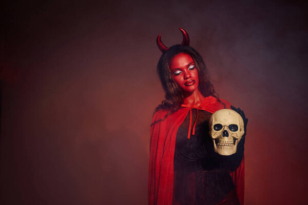A captivating woman in a devil costume stands confidently, clutching a skull in a spooky atmosphere.