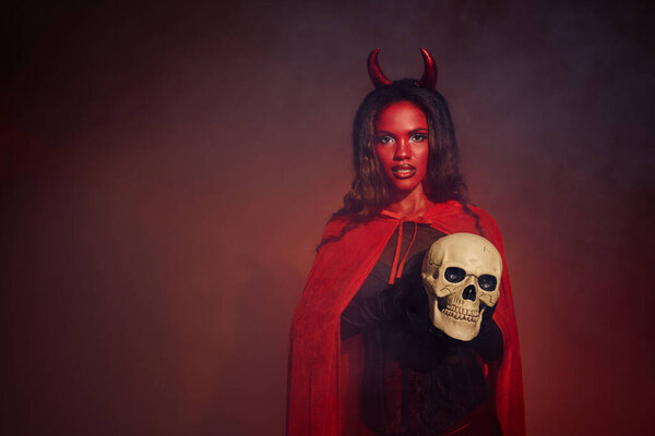 A woman in a bold devil costume holds a skull, radiating Halloween spirit.