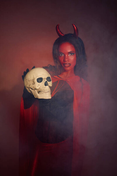 A stunning woman dressed as a devil holds a skull, embraced by mysterious fog and shadows.