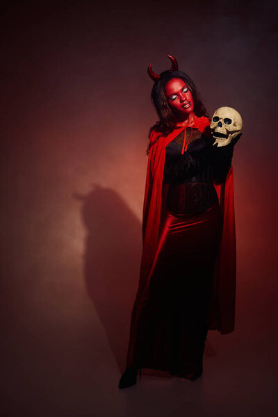 A captivating woman dressed as a devil holds a skull, showcasing Halloween elegance and creativity.
