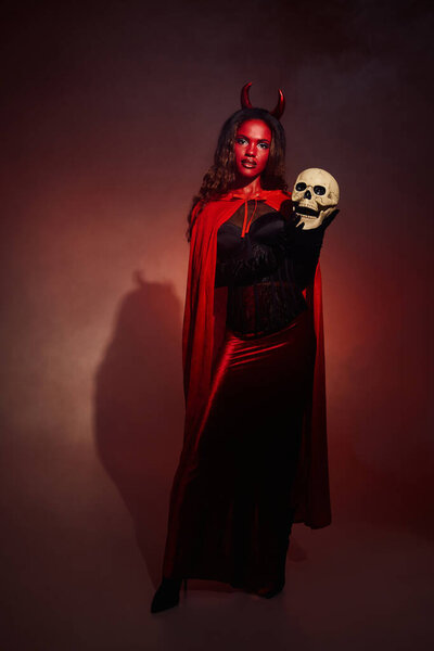 A captivating woman showcases her Halloween spirit in a vibrant devil costume, holding a skull.