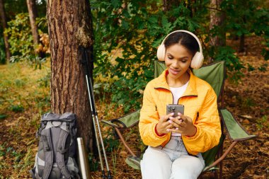 Genç bir kadın, sonbahar güzelliğini kucaklarken telefonuyla müzik dinlerken bir orman kampında dinleniyor..