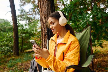 A young African American woman relaxes in the forest, wearing headphones and enjoying her music during autumn. clipart