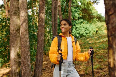 A young African American woman enjoys hiking through a colorful forest as autumn leaves fall around her. clipart