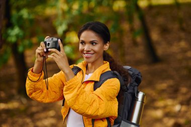 A joyful young woman captures her hiking adventure in a colorful forest during autumn. clipart