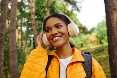 A young African American woman smiles while hiking through a vibrant autumn forest, listening to music. clipart