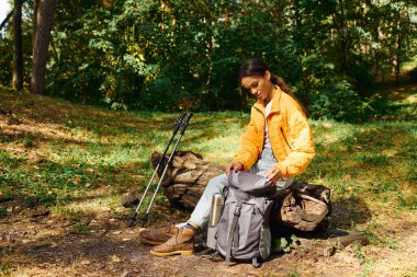 A determined young woman explores a vibrant forest, adjusting her gear amidst the autumn foliage. clipart