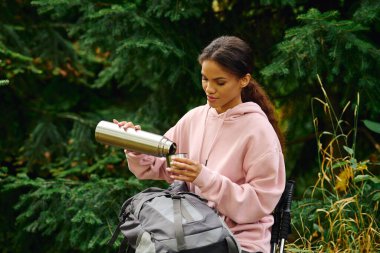 A young woman pauses her hiking adventure to pour a warm beverage, surrounded by vibrant autumn foliage. clipart