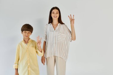 İşitme engelli küçük bir çocuk annesinin yanında poz veriyor. Işıldıyor ve barış işareti yapıyor..