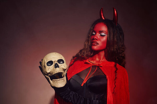 Dressed as a devil, a beautiful woman poses with a skull, radiating Halloween charm and allure.