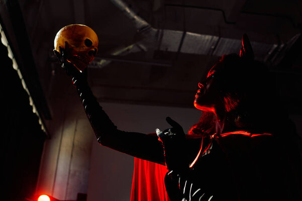 A striking woman dressed in a Halloween costume holds a skull, backlit by a mysterious glow.