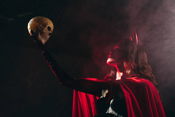 A captivating woman in a Halloween costume raises a skull amidst a foggy, dramatic setting.
