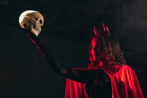 A woman in a red cape and devil horns raises a skull, capturing Halloweens spirit.