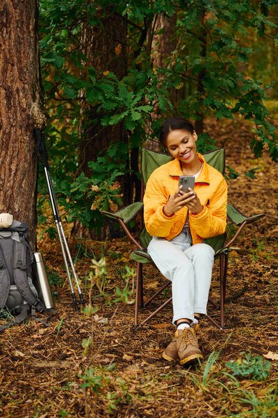 A young woman sits in a chair, smiling at her phone amidst the colorful autumn leaves in the forest.