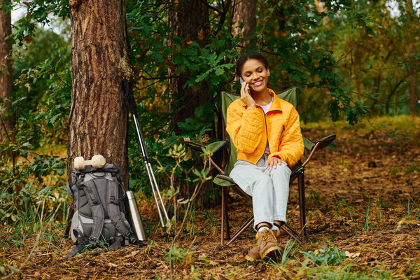 A young woman in an orange jacket is happily chatting on her phone amidst colorful autumn trees while hiking.