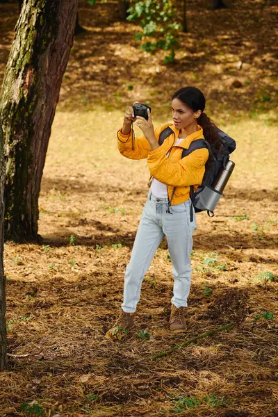 stock image A young African American woman hikes through a vibrant autumn forest, capturing stunning moments with her camera.